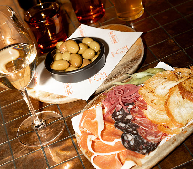An assortment of spanish cured meats, with a bowl of olives and drinks in the background.
