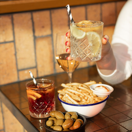 A hand holding a cocktail, with an assortment of other drinks and snacks on the table in the background.